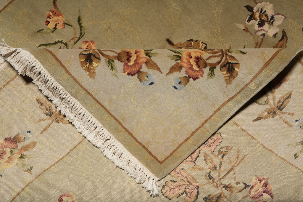 A close-up of a beige woven textile with a floral pattern in muted pink, brown, and green tones, finished with white fringe along one corner.