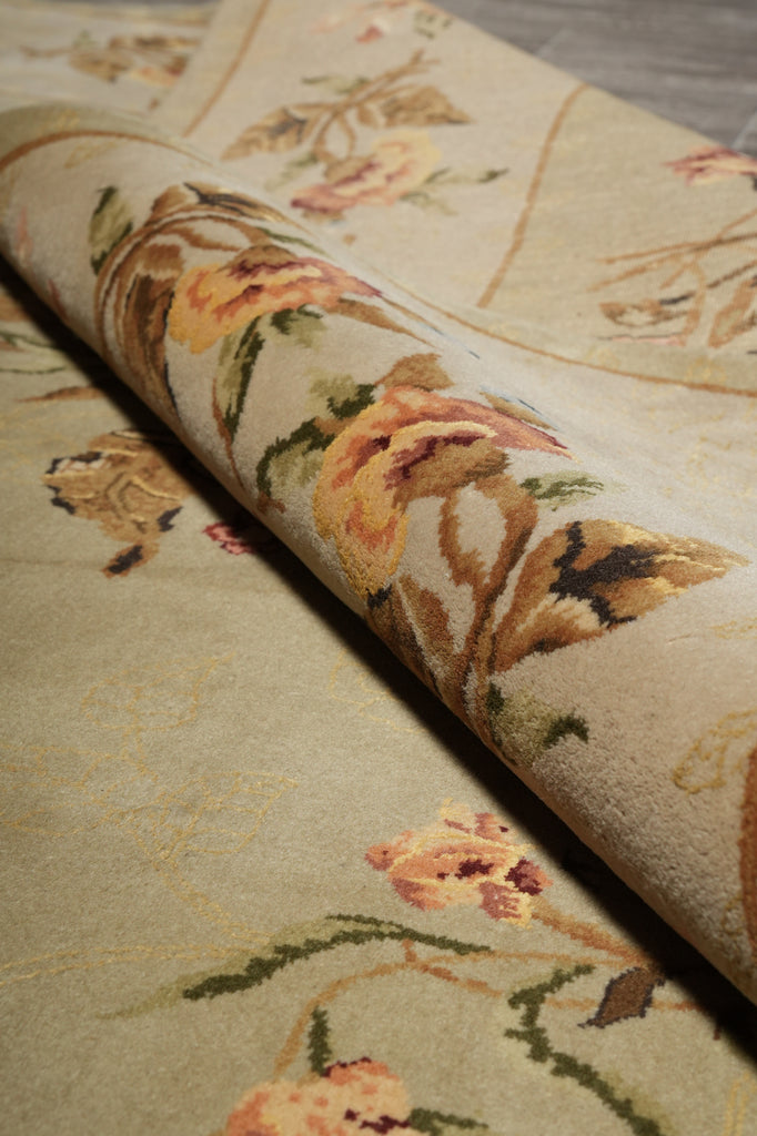 A close-up of a rolled area rug showing a vintage floral pattern in muted tones of beige, gold, green, and brown.