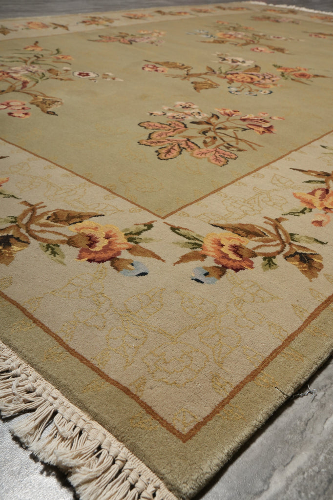 A close-up of a beige area rug with a traditional floral pattern in muted orange, pink, and green tones, featuring a cream-colored fringed edge.