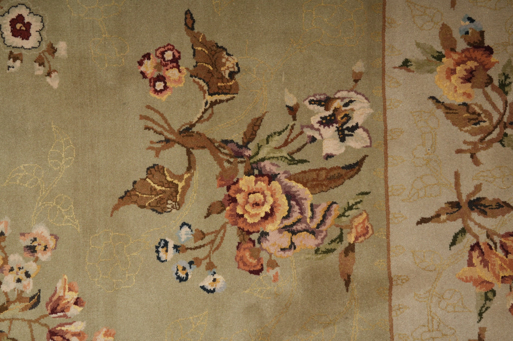 Close-up of a woven textile or rug with a floral pattern on a muted beige background, featuring flowers and leaves in tan, brown, pink, cream, and small blue accents.