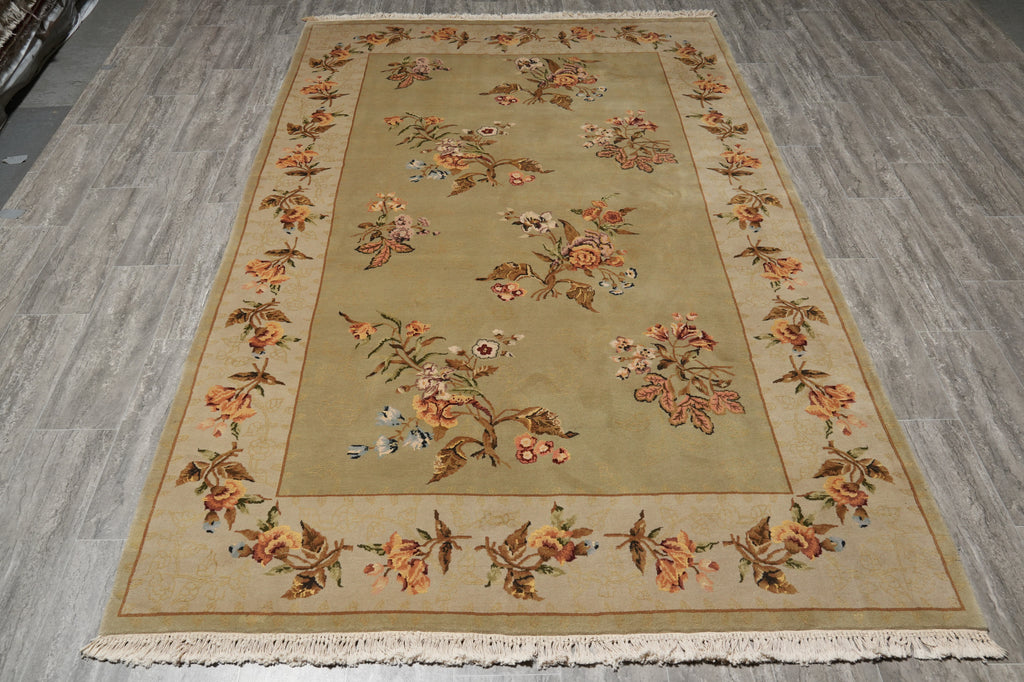A rectangular area rug with a beige and ochre center field, featuring a floral border and small bird motifs, laid out on a gray wood-look floor.