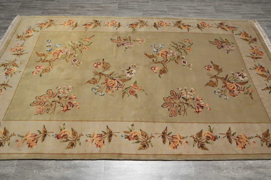 A rectangular vintage-style area rug with a beige center and border, decorated with a floral pattern in muted orange, brown, and blue tones, laid out on a gray tiled floor.