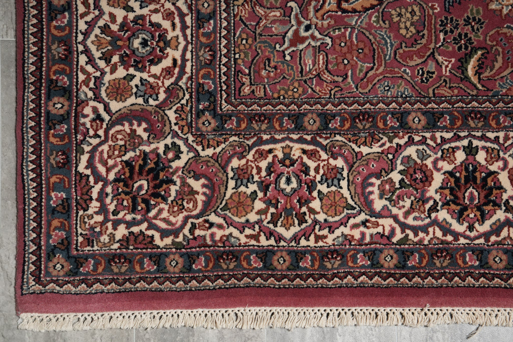 A close-up of an ornate traditional area rug corner, showing detailed floral and vine motifs in deep red, beige, navy blue, and rust tones, with cream fringe visible at the bottom.