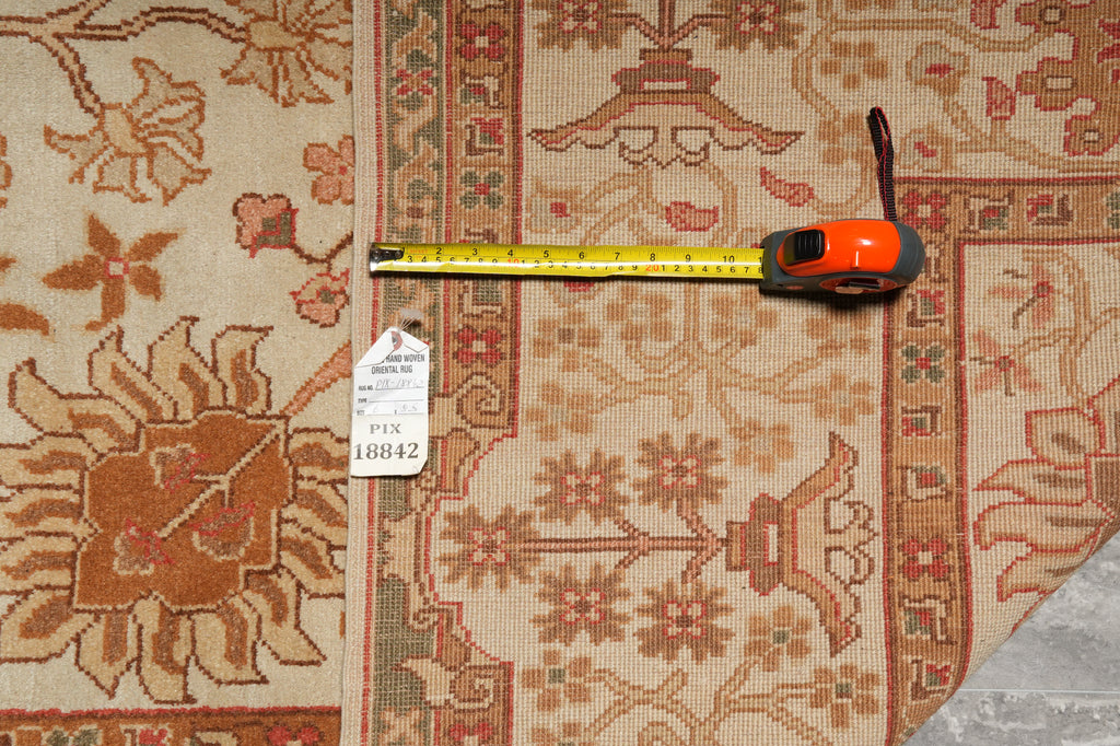 A patterned area rug in beige, rust, and terracotta tones is shown with a yellow tape measure and a white inventory tag laid across it to indicate size.