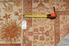 Load image into Gallery viewer, A patterned area rug in beige, rust, and terracotta tones is shown with a yellow tape measure and a white inventory tag laid across it to indicate size.