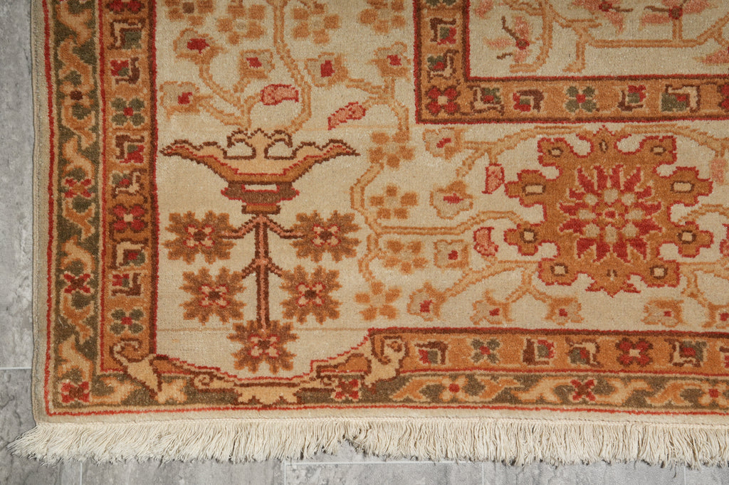 Close-up of a traditional patterned rug corner showing cream, rust, and olive green motifs with fringe along the bottom edge.
