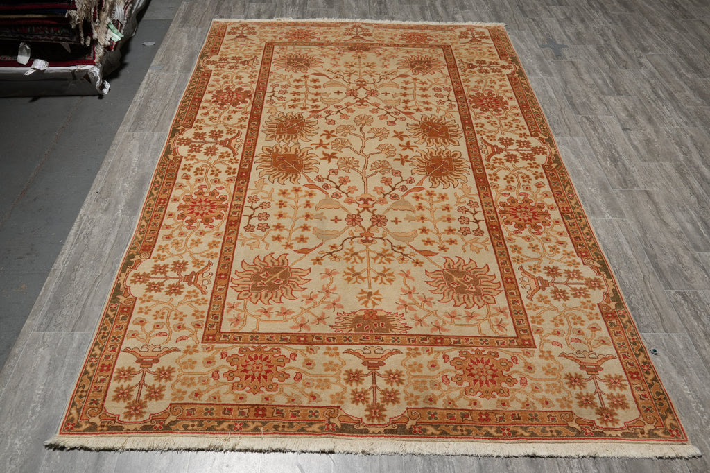 A rectangular area rug with an intricate floral and geometric pattern in beige, rust red, and orange tones, laid out on a gray wood-plank floor.