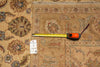 Load image into Gallery viewer, A square patterned area rug in beige and brown tones with floral motifs, shown with a yellow tape measure and a white size tag on top.