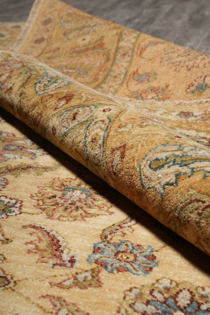 A close-up of a rolled area rug showing its detailed traditional pattern in warm beige, gold, blue, and red tones.
