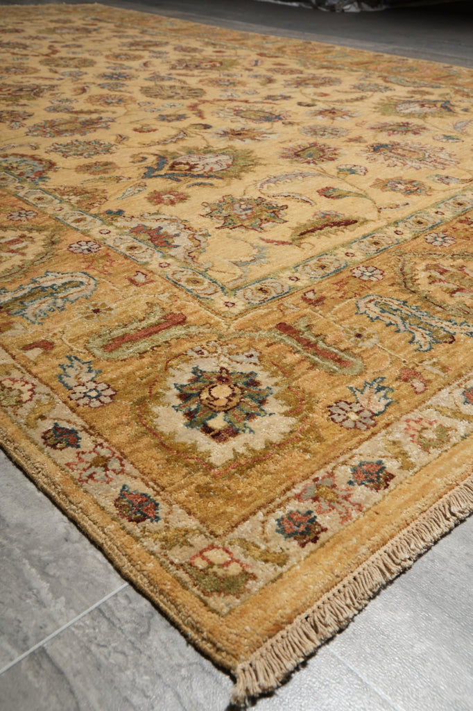 A close-up view of an ornate traditional area rug with a warm beige and gold base, featuring intricate floral and geometric patterns in muted red, green, and blue, with a fringed edge visible on the left against a gray tiled floor.