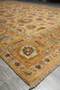 Load image into Gallery viewer, A close-up view of an ornate traditional area rug with a warm beige and gold base, featuring intricate floral and geometric patterns in muted red, green, and blue, with a fringed edge visible on the left against a gray tiled floor.