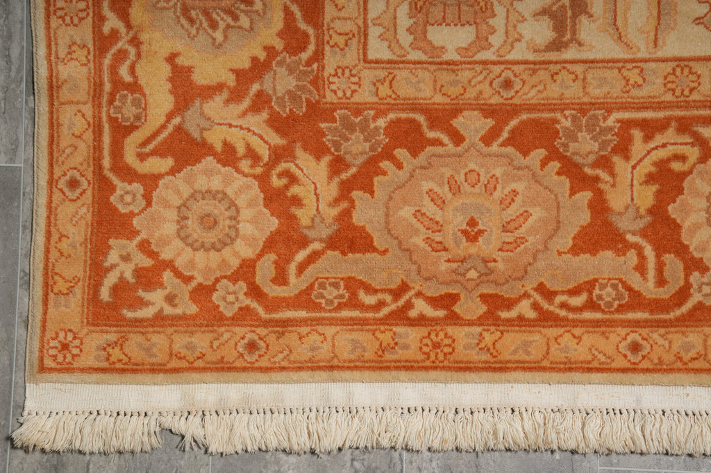 Close-up of an ornate area rug corner showing a warm orange and beige floral medallion pattern with cream fringe at the bottom, laid on a gray floor.