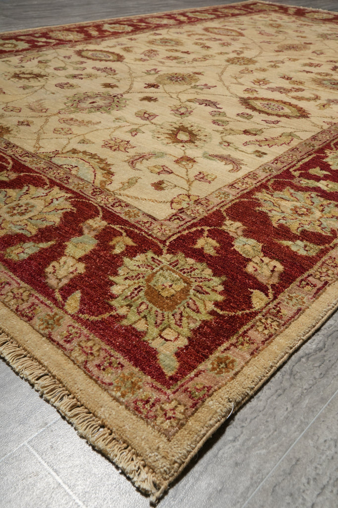 A traditional oriental-style area rug with a beige field and deep red borders, featuring floral motifs and fringed edges, laid out on a gray tiled floor.