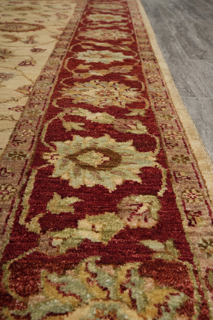 A close-up view of a traditional area rug with a deep red central field, beige floral motifs, and green accents, laid out on a gray floor.