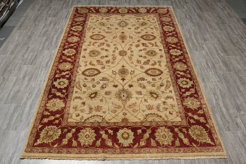A rectangular traditional area rug with an ornate floral pattern, featuring a beige center field, deep red borders, and gold accents, laid out on a gray wood-look floor.