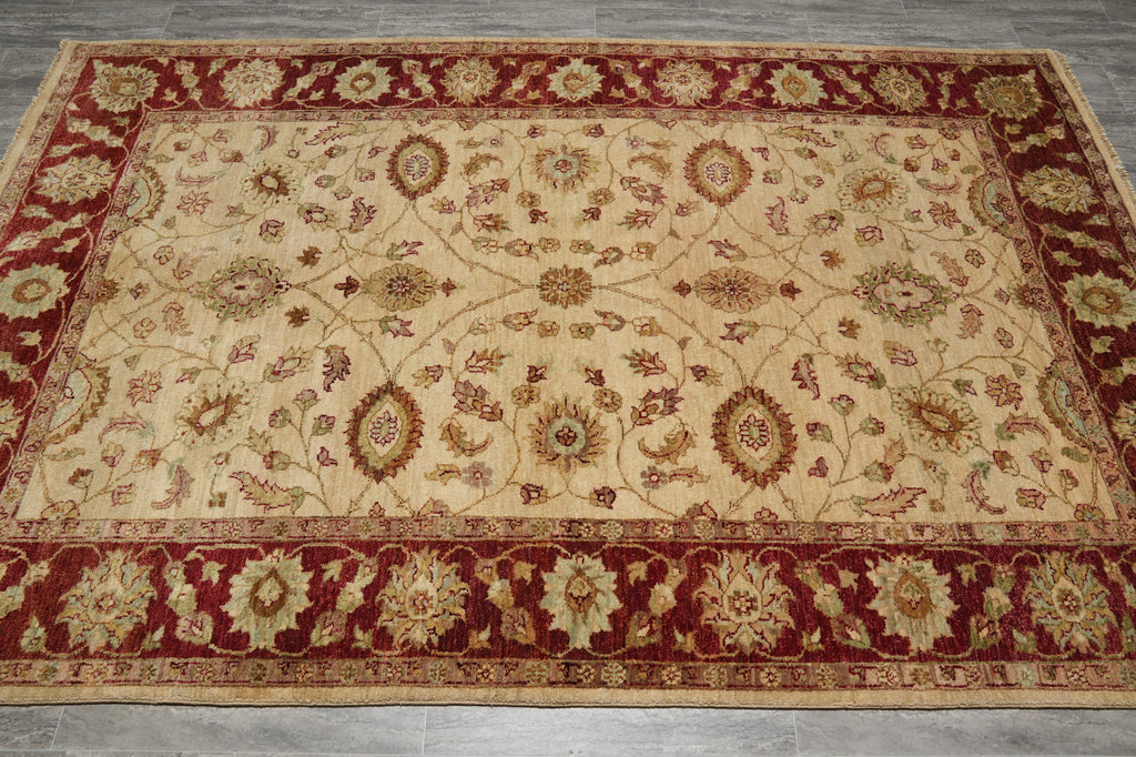 A rectangular area rug with an ornate floral and medallion pattern, featuring a cream center field, deep red borders, and muted green accents, laid out on a gray tiled floor.