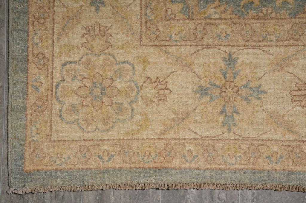 Close-up of an area rug corner showing a beige and tan field with blue and muted green floral medallions, a patterned border, and fringed edge over gray flooring.