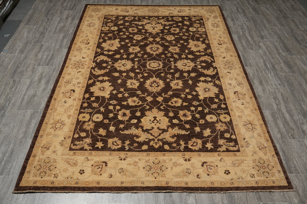 A rectangular area rug with an ornate floral pattern, featuring a dark brown center, a beige inner border, and a darker brown outer border, shown laid out on gray wood-look floor tiles.