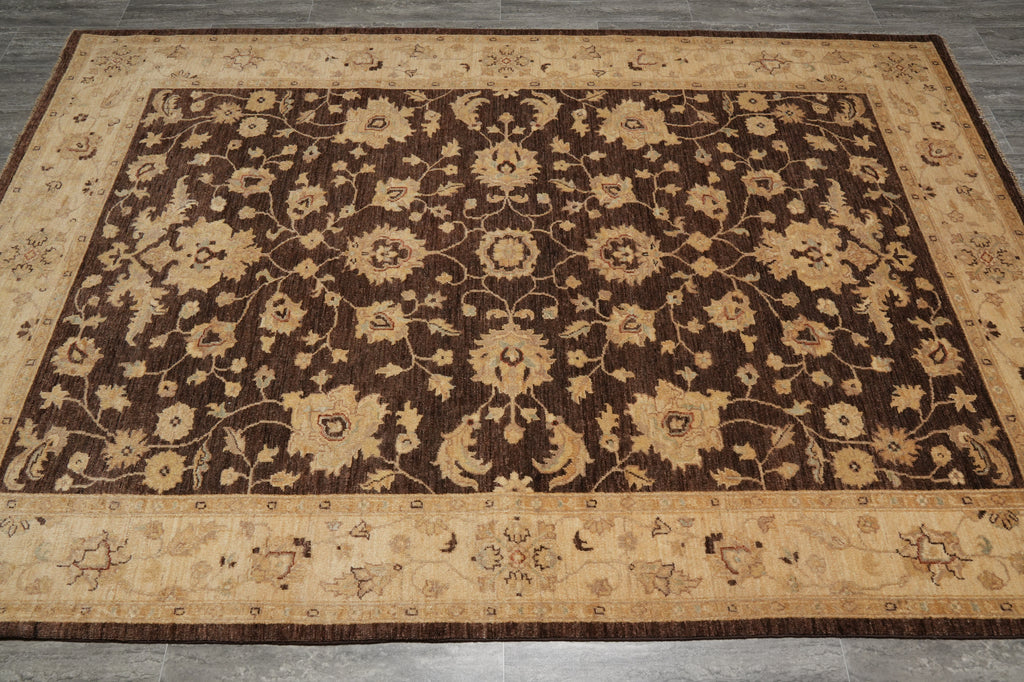 A rectangular area rug with an ornate floral and vine pattern, featuring a dark brown center field, beige borders, and small bird motifs along the lower border, shown on a gray tiled floor.
