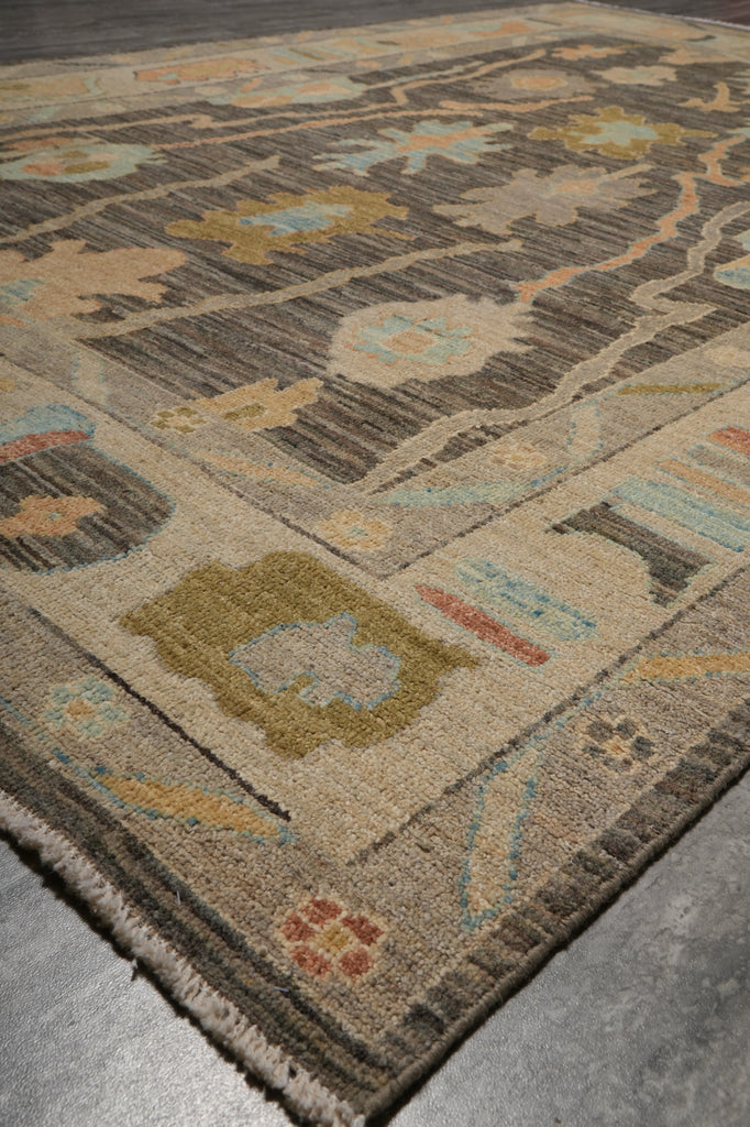 A close-up view of a traditional area rug with a geometric and floral pattern in beige, brown, muted blue, and olive green, shown on a gray concrete floor.