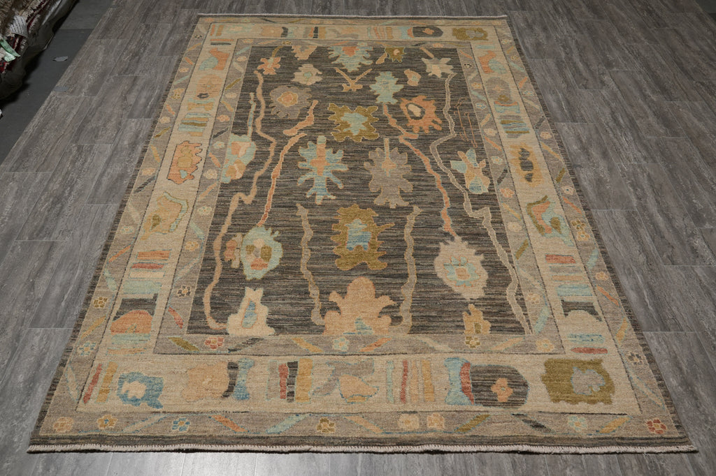 A rectangular area rug with an intricate traditional pattern, displayed on a dark gray wood-look floor. The rug features a dark charcoal center with beige, light blue, and ochre accents, surrounded by a wide beige border.
