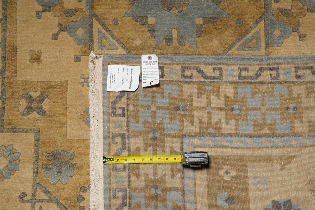 A close-up of a patterned area rug corner with a tape measure and tags laid on top. The rug has a beige and tan base with gray and muted blue geometric motifs and a cream fringe edge.