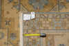 Load image into Gallery viewer, A close-up of a patterned area rug corner with a tape measure and tags laid on top. The rug has a beige and tan base with gray and muted blue geometric motifs and a cream fringe edge.