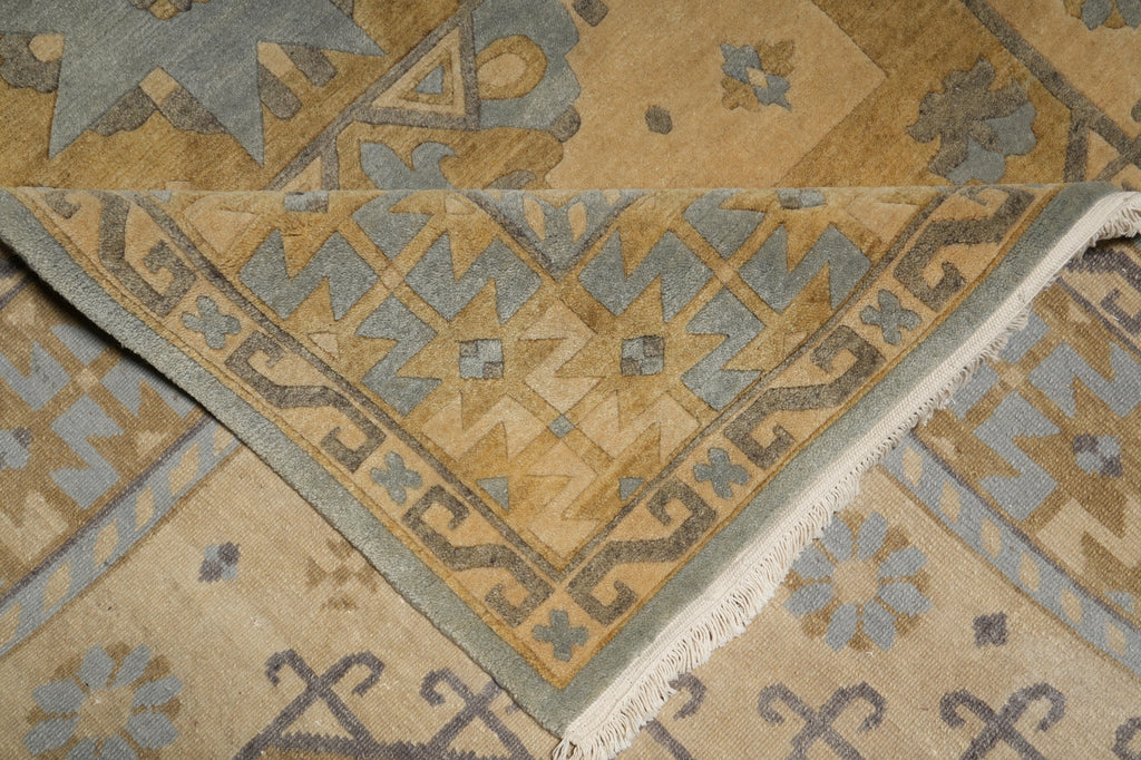 Close-up of a patterned area rug showing a folded corner that reveals the cream fringe and detailed geometric motifs in tan, beige, and muted blue-gray.