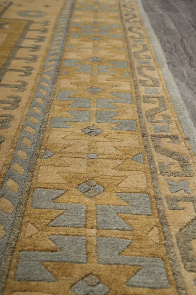 A close-up view of a patterned area rug with a warm tan and beige base, featuring geometric motifs in muted gray and a border in a taupe-brown tone.