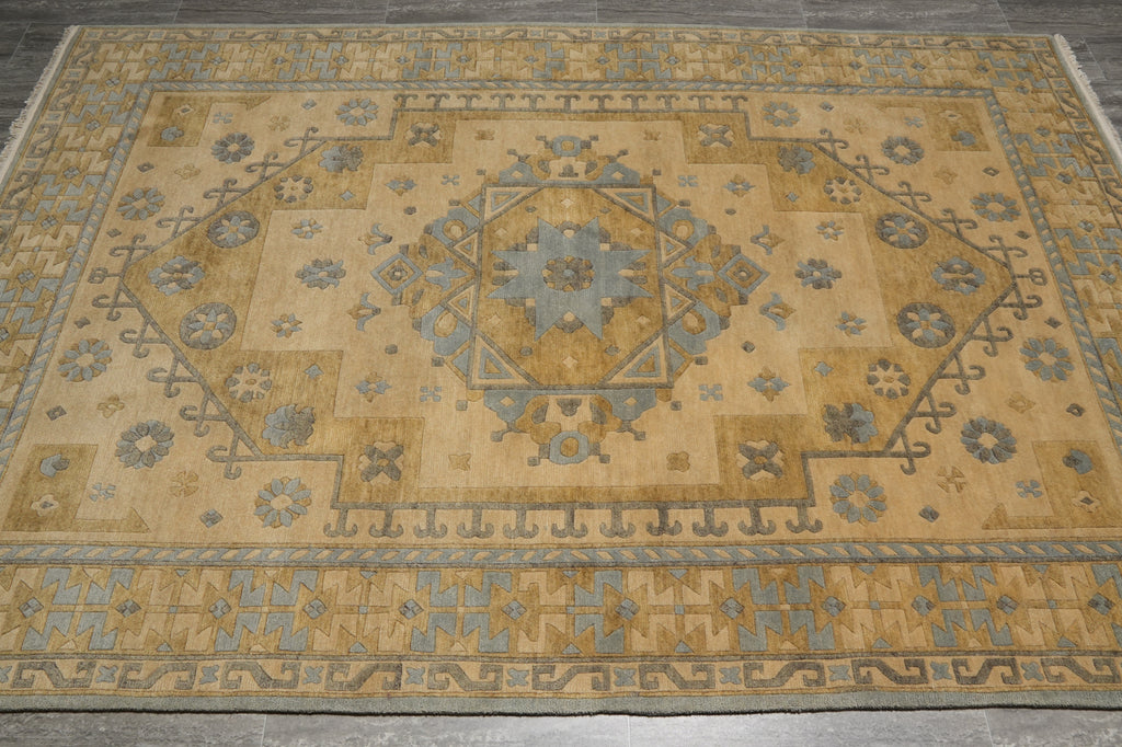 A rectangular area rug with a traditional medallion pattern in warm beige, muted gold, and dusty blue-gray, shown from a top-down perspective on a dark gray floor.