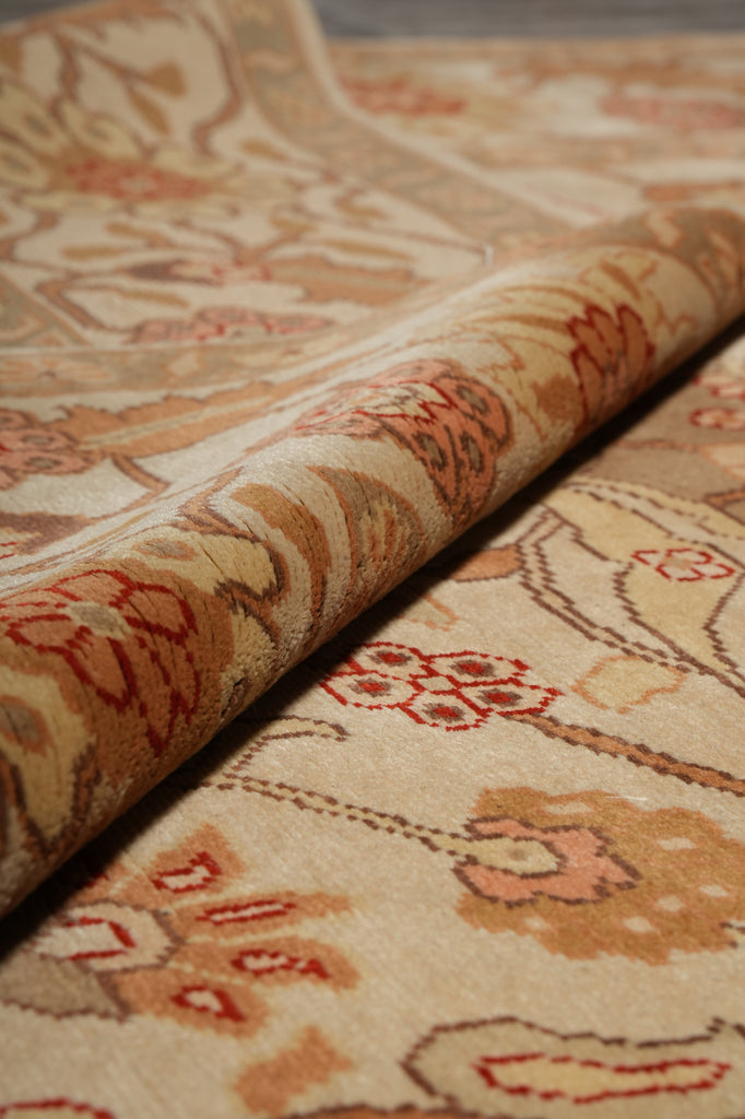 A close-up of a patterned area rug with a rolled section showing its texture and design. The rug features a traditional floral motif in beige, brown, and muted red tones.