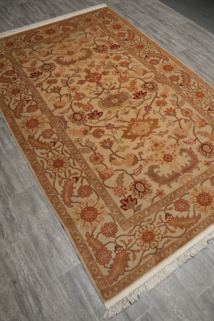 A rectangular traditional area rug with an intricate floral and vine pattern, displayed on a gray wood-look floor. The rug features a warm beige base with rust red, brown, and cream details, complete with white fringe on two short ends.