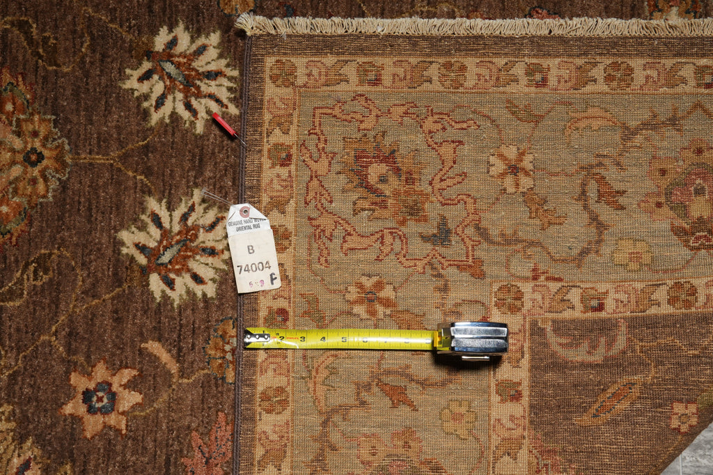 A close-up of a patterned area rug with a yellow tape measure and a white tag placed on it for scale, showing the rug's intricate floral and geometric designs in brown, beige, and rust tones.
