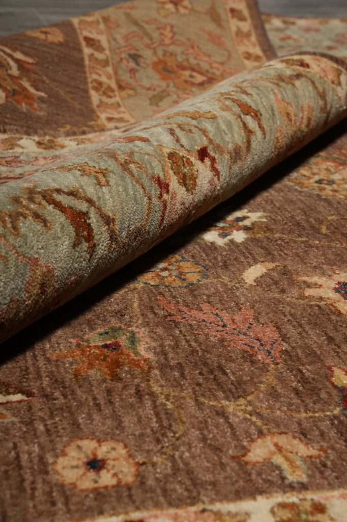 A close-up of a traditional patterned area rug, showing a rolled section that reveals the underside in a muted sage green with rust and beige motifs, while the top surface displays a warm brown field with floral and vine designs.