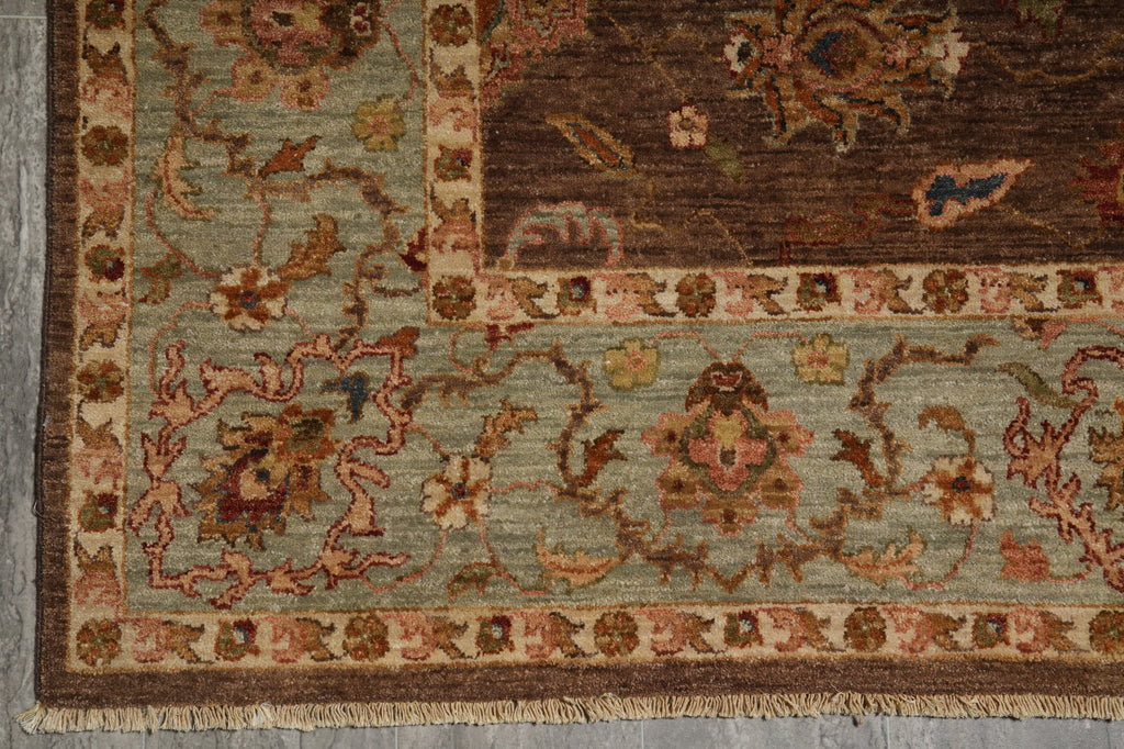 Close-up of an ornate patterned rug corner showing a muted sage green field, dark brown border, and floral motifs in beige, rust, and blue.