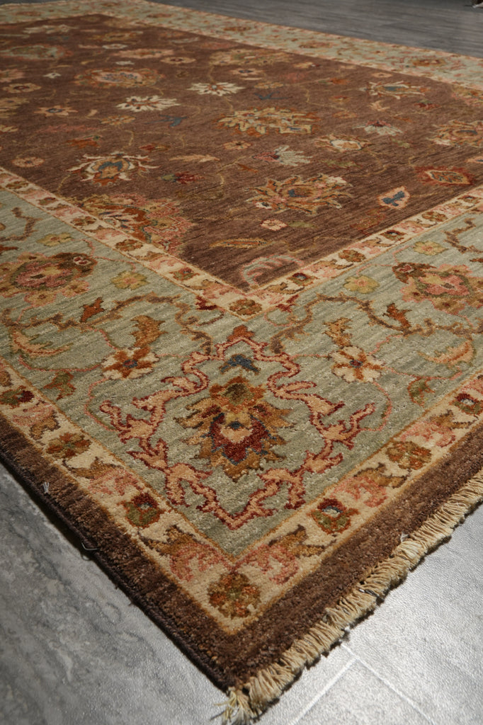 A traditional oriental area rug with a brown central field, a beige and rust border, and a light sage green corner panel, shown on a gray tiled floor.
