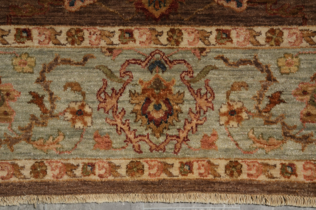 A close-up of an ornate patterned rug showing a central floral medallion and scrolling vine motifs in muted tones of sage green, rust red, beige, and brown, with a fringed edge at the bottom.