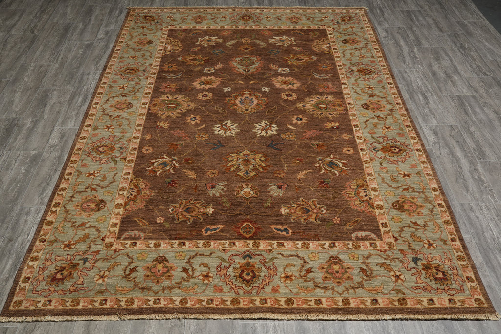 A traditional oriental area rug with a dark brown central field, beige and light blue-green borders, and intricate floral motifs, shown laid out on a gray wood-look floor.