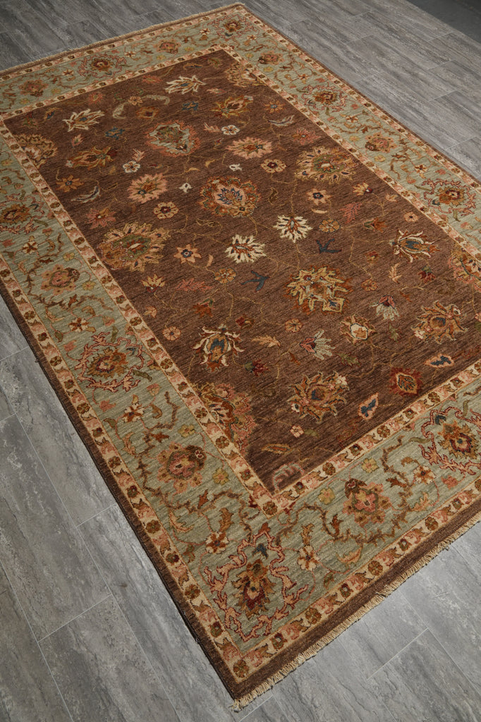A rectangular area rug with an ornate floral pattern, featuring a dark brown central field and a light sage green border, laid out on a gray wood-look floor.