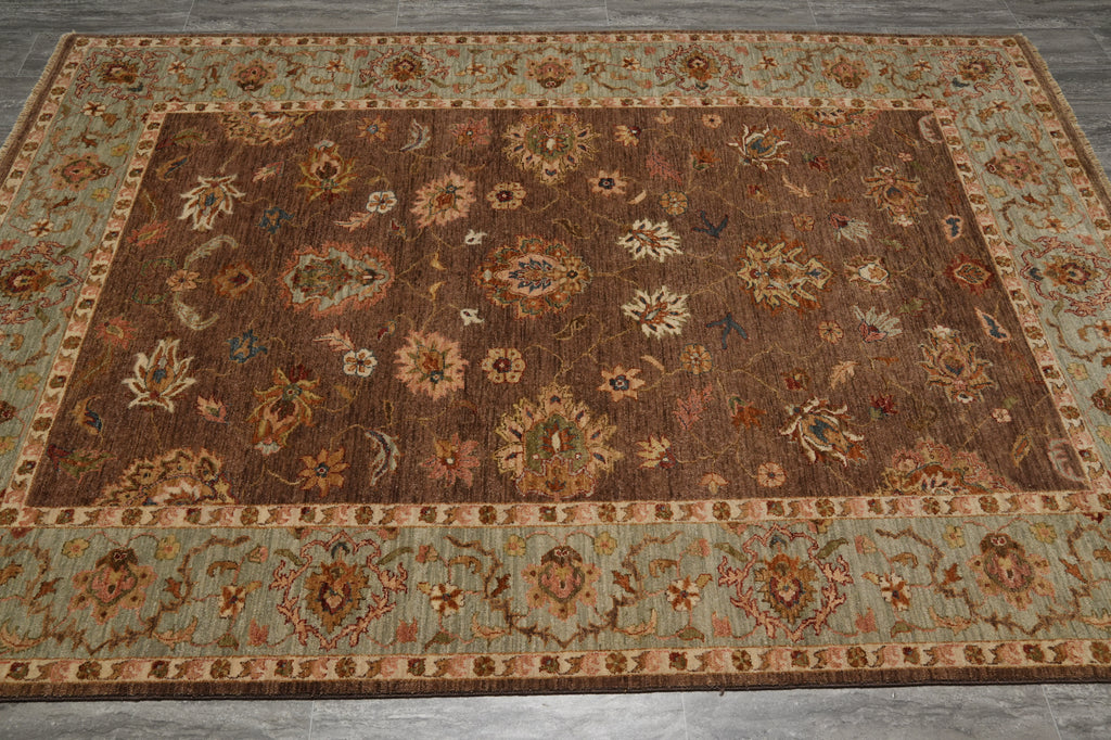 A rectangular area rug with a traditional floral pattern, featuring a dark brown center field and a light sage green border, laid out on a gray tiled floor.