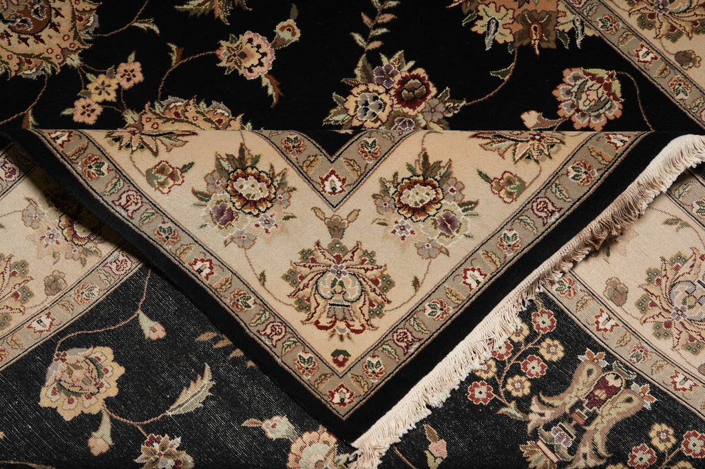 A close-up of a traditional oriental rug with a black field and a cream-colored V-shaped corner section featuring intricate floral and vine patterns in beige, green, and red.