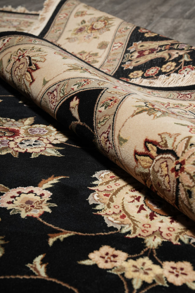 A close-up of a traditional oriental-style rug with a black base and beige floral motifs, partially rolled to show the pattern and texture.