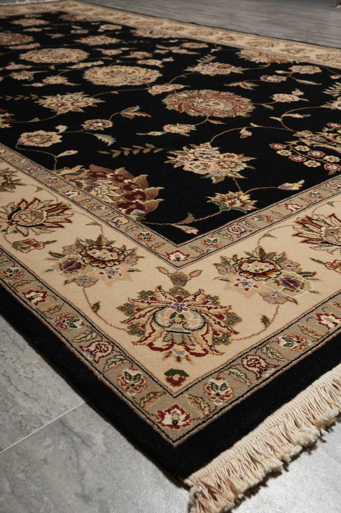 A traditional oriental area rug with a black field, beige borders, and floral motifs in cream, rust, and green, shown on a gray tiled floor.
