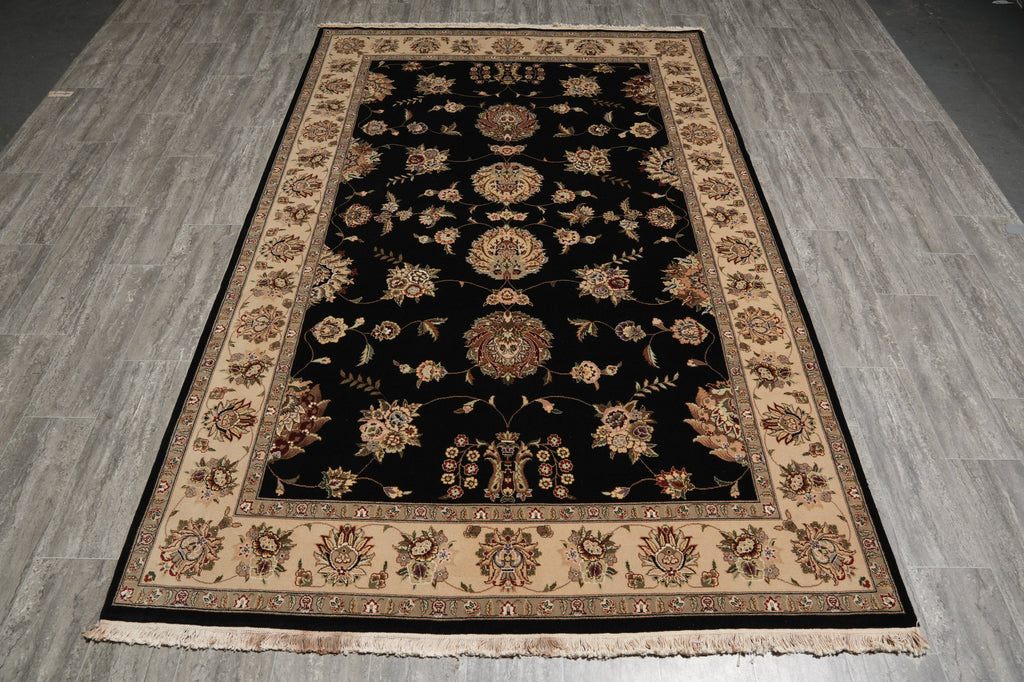 A rectangular area rug with a black center field and beige floral motifs, laid out on a gray wood-look floor.