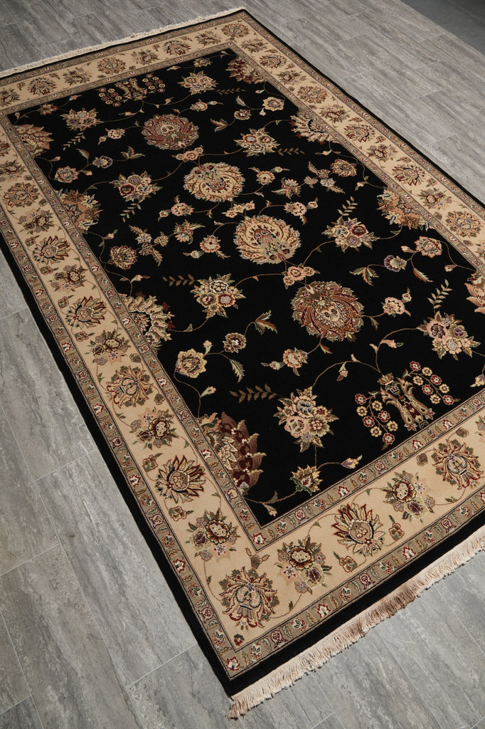 A rectangular area rug with a black field and ornate beige floral border, photographed from above on a gray wood floor.