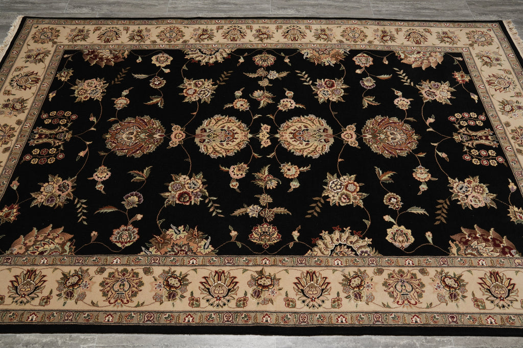 A rectangular area rug with a black center field and ornate floral motifs in beige, brown, and muted red, surrounded by a detailed beige border.