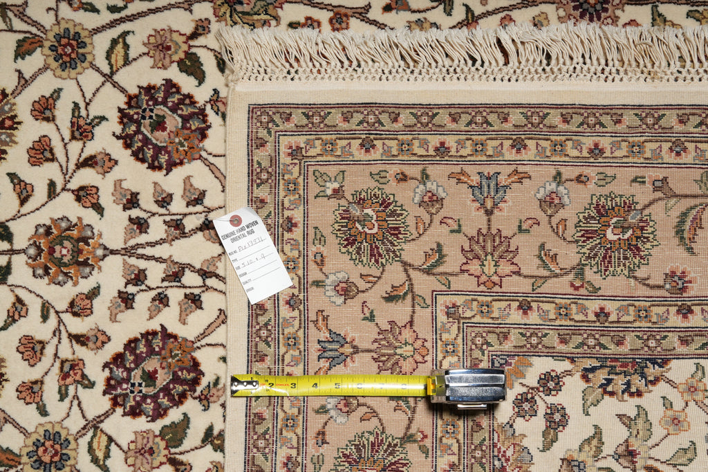 A close-up of a patterned area rug with a cream base and floral motifs in beige, brown, and muted red tones. A yellow tape measure lies across the rug, and a white product tag is attached near the left side.