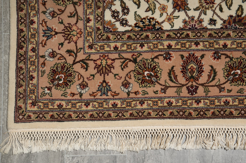 A close-up of a traditional patterned rug corner showing cream fringe, a dusty pink field, and ornate floral motifs in beige, brown, and muted red on a gray floor.