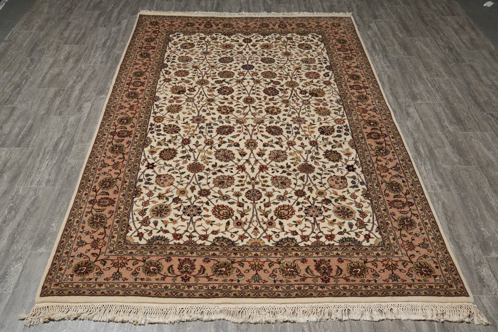An oriental-style area rug with an intricate floral pattern, laid flat on a gray wood-look floor. The rug features a cream center field, a dusty rose border, and dark brown detailing, complete with a fringed edge.
