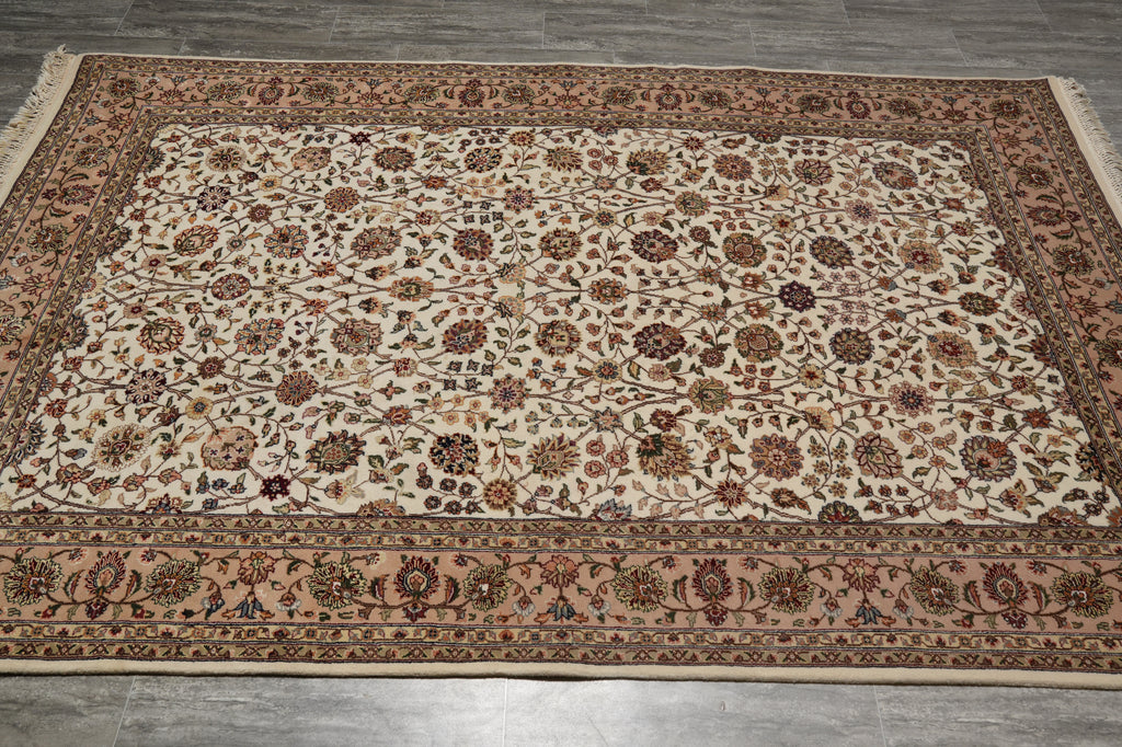An oriental-style area rug with an intricate floral pattern, laid out on a gray tiled floor. The rug features a cream-colored center field, a dusty rose border, and dark brown and green details throughout the design.